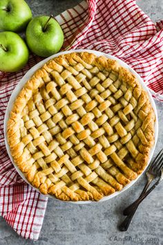 an apple pie sitting on top of a red and white checkered cloth next to two green apples