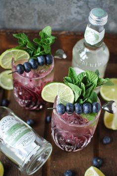 two glasses filled with blueberries and limes on top of a wooden table next to bottles