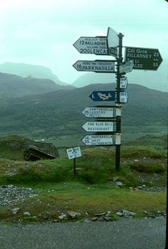 a pole with many signs on it in front of some hills and grass near a road