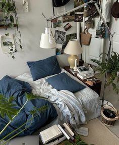 an unmade bed with blue sheets and pillows in a room filled with books, plants and pictures on the wall