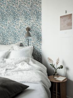 a white bed sitting next to a wall covered in blue and white wallpaper on top of a wooden night stand