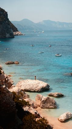 a man is standing on the beach near some rocks and boats in the blue water