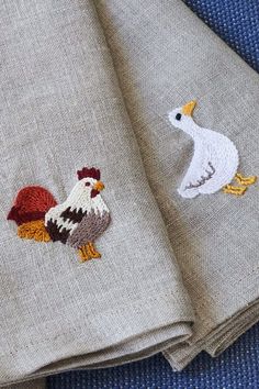 two pieces of linen with embroidered roosters on them, one in grey and the other in red