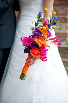 a bride and groom standing next to each other in front of a brick wall holding a colorful bouquet