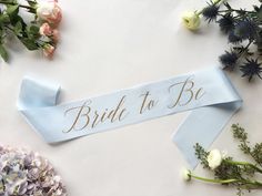 the bride to be sash is laying on top of some flowers and blue ribbon with gold lettering