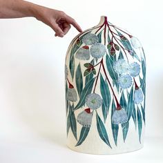 a hand is reaching into a vase with blue flowers on it and green leaves painted on the bottom