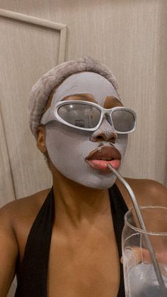 a woman in a black tank top drinking from a glass with a silver mask on her face