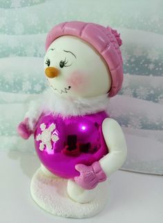 a snowman figurine holding a christmas ornament in its hands and wearing a pink hat