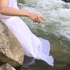 a woman in white dress sitting on rock pointing at river with rocks and water behind her