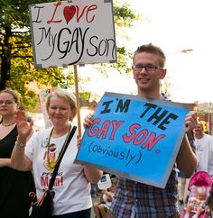 a man holding a sign that says i'm the gay son obviously with other people behind him