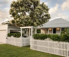 a white picket fence in front of a house with trees and bushes on the lawn