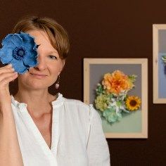 a woman holding up a blue flower in front of her face with three pictures on the wall behind her