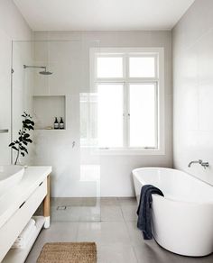 a white bath tub sitting under a window next to a sink in a bathroom with a rug on the floor