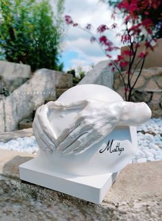 a sculpture of two hands holding each other in front of some rocks and flowers on the ground