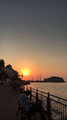 the sun is setting over the water and people are sitting on benches