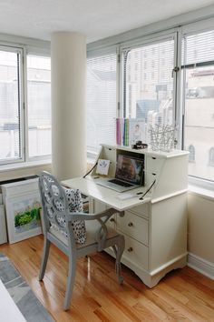 a desk with a computer on top of it in front of two windows and a wooden floor