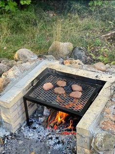 an outdoor fire pit with hamburgers cooking on it's grill, in front of some rocks and grass