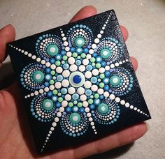 a hand is holding a small square painted with dots and circles on black, blue, green and white colors