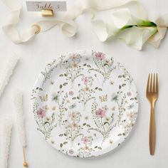 a white plate with flowers on it next to gold forks and napkins