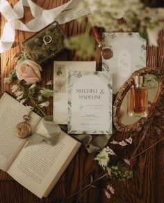 an open book and some flowers on top of a wooden table next to two wedding rings