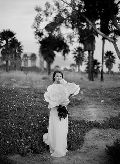 black and white photograph of woman in dress standing on dirt path with palm trees behind her