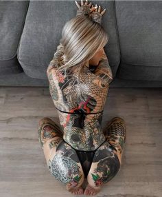 a woman with tattoos sitting on the floor in front of a gray couch and holding her hands behind her head