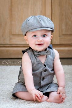 a baby sitting on the floor wearing a gray suit and hat, smiling at the camera