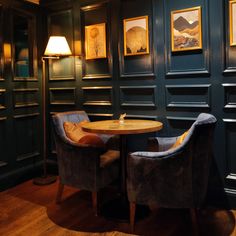 two chairs sitting in front of a table with a lamp on it next to some framed pictures