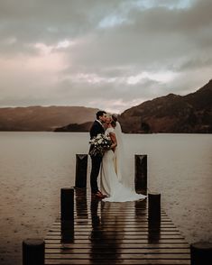 a bride and groom kissing on a dock in the middle of a lake with mountains in the background