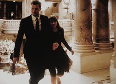 a man and woman dressed in black walking down a hall with columns on either side