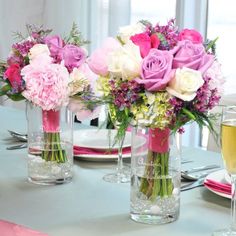 pink and white flowers are in vases on a table with wine glasses, plates and utensils