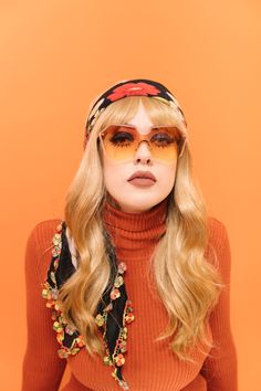 a woman with long blonde hair wearing sunglasses and a headband on an orange background