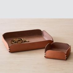 two brown leather trays with handles on a wooden table top, one holding a gold ring and the other is empty