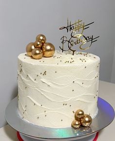 a white and gold birthday cake on a silver platter with two golden balls next to it