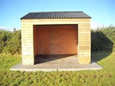 an empty wooden garage in the middle of a field with grass and bushes around it