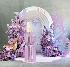 there is a cake on top of a stand with purple flowers and butterflies around it