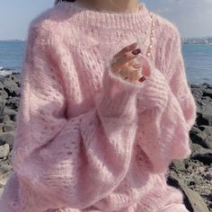 a woman in pink sweater sitting on rocks by the ocean with her hand up to her face