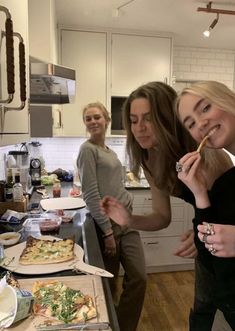 three women standing in a kitchen with pizzas on the counter and one is eating