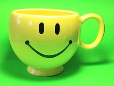 a yellow coffee cup with a smiley face painted on the front and sides, sitting on a green surface