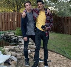 two young men standing next to each other in front of a pond and wooden fence