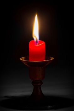 a red candle is lit on a small wooden stand in the dark with its bright flame