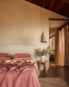 a bed with pink sheets and pillows next to a wooden table in front of a window