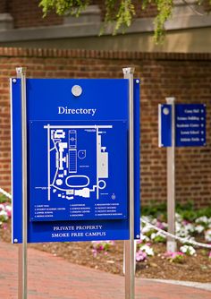 a blue sign sitting on the side of a road next to a brick building and flowers