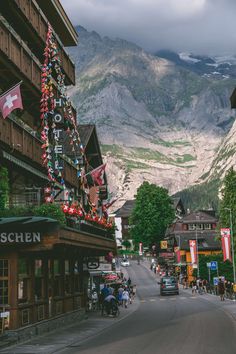 a town with mountains in the background and people walking on the sidewalk near buildings that have flags hanging from them