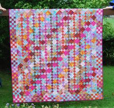 a person holding up a colorful quilt in front of some bushes and trees with flowers