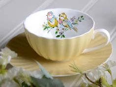 a yellow cup with two birds painted on the side sitting on a saucer next to flowers