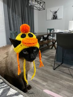 a siamese cat wearing a knitted hat with an orange and yellow bird on it's head