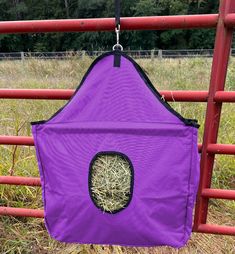 a purple bag with hay in it hanging on a red fence near some grass and trees