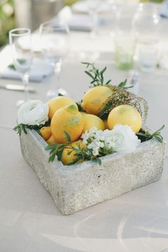 an arrangement of lemons and white flowers in a stone bowl on a table with wine glasses