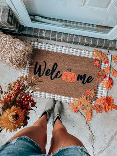 a welcome mat with the words welcome on it and fall leaves around it, next to a woman's legs